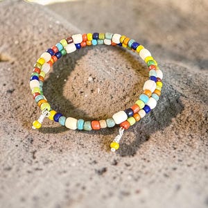 May include: A colorful beaded bracelet with a spiral design. The bracelet features small, square beads in various colors, including white, blue, yellow, orange, and green. The bracelet is resting on a textured, gray surface.