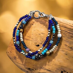 May include: A multi-strand beaded bracelet featuring a mix of blue, turquoise, silver, and metallic beads. The bracelet is secured with a silver-toned circular toggle clasp and lobster claw closure, showcasing a variety of bead shapes and textures.