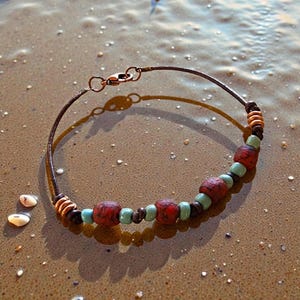 May include: Handmade bracelet with a dark brown wire and copper clasp. The bracelet features red, turquoise, and black beads. The bracelet is resting on a sandy surface, with seashells in the background.