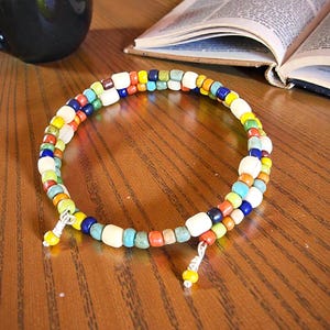 May include: A colorful beaded bracelet with a spiral design. The bracelet features a variety of small, square beads in shades of white, yellow, orange, red, green, blue, and brown. The bracelet is resting on a wooden surface.
