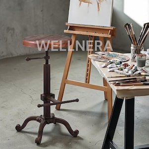 May include: A brown adjustable artist's stool with a round wooden seat and a dark metal base. The word "WELLNERA" is printed on the seat. An easel, paint supplies, and a table are in the background.