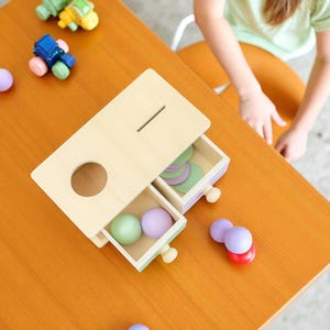 May include: Wooden educational toy with a light wood finish. The toy has a slot, a large hole, and two drawers containing colorful balls and discs. Small toy cars are scattered nearby. The toy is on an orange table.