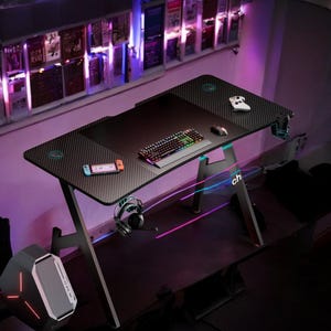 May include: Black gaming desk with a carbon fiber texture, featuring a keyboard, mouse, and game controllers. The desk has a headset holder and a computer tower. The desk is approximately 60 inches long.