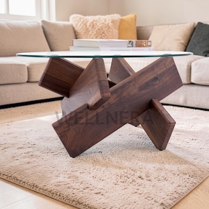 May include: A modern coffee table with a clear glass top and a dark brown, geometric wooden base. The base is constructed from intersecting wooden beams. The table is placed on a beige rug in a living room setting.
