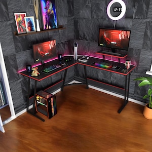 May include: An L-shaped black gaming desk with red trim and LED lighting. The desk has two computer monitors, a keyboard, and a mouse. A gaming PC, framed artwork, and a digital clock are also visible.