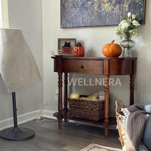 May include: A dark wood console table with a single drawer and lower shelf, featuring turned legs. On top sits a framed black cat illustration, a small orange pumpkin, and a larger orange pumpkin next to a green glass vase with white flowers.