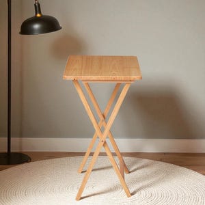 May include: A light-colored wooden folding table with an X-shaped base and a rectangular top. The table is made of natural wood and is set on a white rug. The table is approximately 24 inches tall.