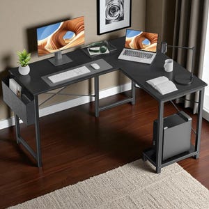 May include: A black L-shaped computer desk with a metal frame. The desk has a monitor, laptop, keyboard, mouse, and a small plant. A computer tower sits on a lower shelf. The desk is on a hardwood floor with a beige rug.