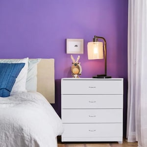 May include: A white four-drawer dresser with silver handles is set against a purple wall. A decorative lamp with a square shade and a small framed picture sit on top of the dresser. A bed with white bedding and blue pillows is in the foreground.