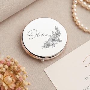 May include: A round, silver compact mirror with a floral design and the name "Olivia" in script. The mirror is set on a neutral surface with a pearl necklace and dried flowers in the background. The compact has a rose gold trim.