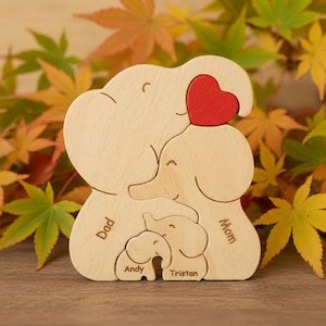 May include: A wooden elephant family puzzle with a red heart. The puzzle includes the words "Dad", "Mom", "Andy", and "Tristan". The puzzle is made of light-colored wood.
