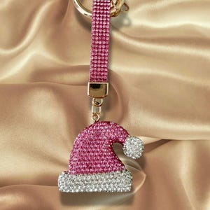 May include: A keychain featuring a gold-tone metal ring attached to a vertical strap covered in pink rhinestones. The strap connects to a charm shaped like a Santa hat, which is encrusted with pink rhinestones and features a white rhinestone trim.