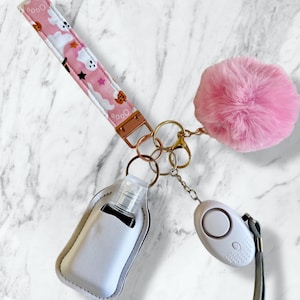May include: A keychain accessory set featuring a pink fabric wristlet with a ghost and pumpkin pattern, a fluffy pink pom-pom, a white holder containing a small clear sanitizer bottle, and a white personal safety alarm with a gray wrist strap.