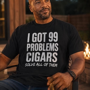 May include: Black t-shirt with the text "I GOT 99 PROBLEMS CIGARS SOLVE ALL OF THEM" in white. The person is wearing blue jeans and a black watch. A lit cigar is held in the right hand.