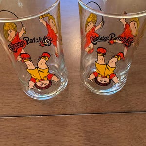 May include: Two clear drinking glasses featuring Cabbage Patch Kids graphics. Each glass has a cartoon image of a child in red and yellow clothing, with the words "Cabbage Patch Kids" in red. The glasses are on a wooden surface.