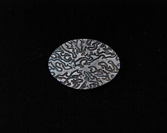 Oval silver pin with a linear pattern, highlighted with blue iridescent patina