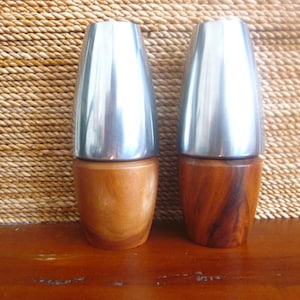 May include: Two salt and pepper shakers with stainless steel tops and wooden bases. The wooden bases have a natural finish, and the shakers are displayed on a wooden surface.