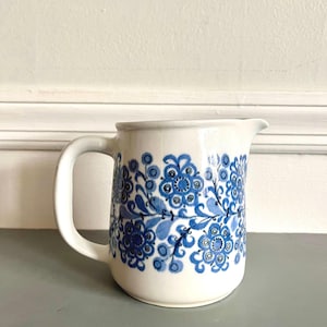 May include: A white ceramic pitcher with a blue floral pattern. The pitcher has a handle and a spout for pouring. The floral design features stylized flowers and leaves in varying shades of blue.