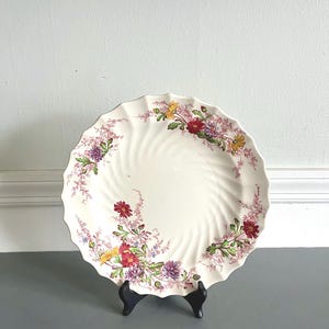 Puede incluir: Un plato de cerámica blanca con borde festoneado y diseño floral. El plato presenta un patrón en espiral y está decorado con flores coloridas en tonos rojos, amarillos y morados. El plato se exhibe sobre un soporte negro.