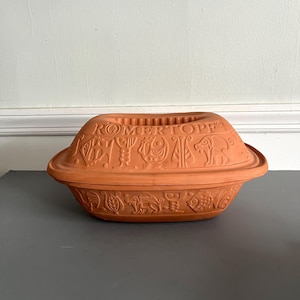Op de afbeelding: Een terracotta Römertopf kleibakker met een deksel. De bakker heeft reliëfontwerpen en het woord "RÖMERTOPE" op het deksel. De bakker staat op een grijze ondergrond.