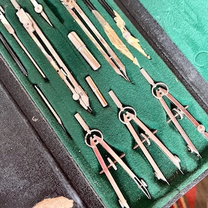 Può includere: Un set da disegno vintage in una custodia nera con interno in velluto verde. Il set include diversi compassi, divisori e altri strumenti da disegno color argento. La custodia è aperta, rivelando gli strumenti ben disposti.