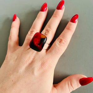 Puede incluir: Un anillo de plata con una piedra grande, rectangular y multicolor. La piedra tiene una mezcla de tonos rojos, negros y amarillos. La mano tiene uñas largas pintadas de rojo. El anillo es una pieza audaz.