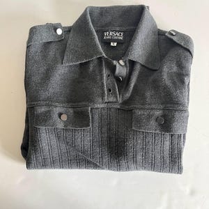 Puede incluir: Una camisa de punto gris con cuello y cierre de botones. La camisa tiene dos bolsillos delanteros con cierres de botón plateados y charreteras en los hombros. La marca "VERSACE JEANS COUTURE" es visible en la etiqueta.