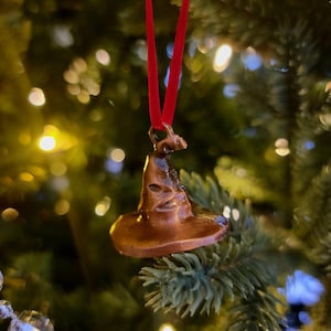 Peut inclure: Ornement de Noël en forme de chapeau de sorcier de couleur bronze, suspendu par un ruban rouge. L'ornement est placé sur un fond de branches de sapin de Noël vertes et de lumières scintillantes, créant une scène de fête.