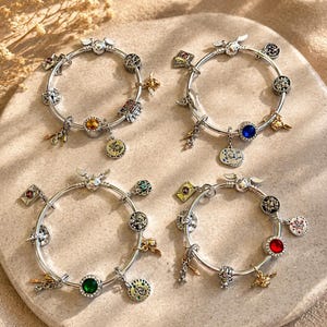 May include: Four silver-toned charm bracelets featuring various miniature pendants, including winged spheres, envelopes, and circular charms with embedded coloured stones in yellow, blue, green, and red, arranged on a circular stone surface.