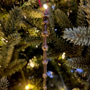 May include: A bronze wand-shaped Christmas tree ornament suspended by a red ribbon. The wand features a detailed design with decorative spheres and a textured handle. The ornament hangs against a blurred backdrop of a Christmas tree with fairy lights.