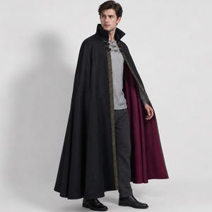 May include: A floor-length black cloak with a high collar, featuring a deep burgundy lining and a decorative gold-toned trim along the front edges. The garment is secured at the neck with two metallic clasps and worn over a grey shirt and trousers.