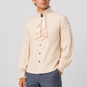 Pode incluir: Uma camisa de manga comprida cor de creme com um jabot franzido na gola alta, preso com um broche de metal escuro. A camisa inclui pregas verticais na frente e nas mangas, com três botões visíveis e punhos nas mangas.