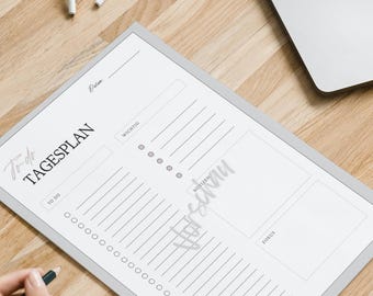 May include: A workspace with a laptop, a black notebook, and a daily planner. The planner has the text "Tagesplan" and a section for notes. A small potted plant and wireless earbuds are also on the wooden desk.