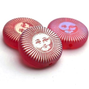 Puede incluir: Tres cuentas redondas rojas con un diseño de cara de sol. Las caras del sol son blancas y moradas claras, con líneas radiales. Las cuentas son translúcidas y tienen un agujero para ensartar.