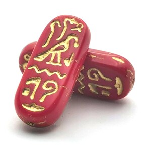 Könnte beinhalten: Zwei rote, ovale Perlen mit goldenen Hieroglyphen-Designs. Die Perlen haben eine leicht abgenutzte, glänzende Oberfläche. Das Design umfasst Vogel- und Wellensymbole. Diese Perlen sind wahrscheinlich für die Schmuckherstellung oder dekorative Zwecke bestimmt.