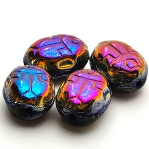 Op de afbeelding: Vier iriserende scarabee kever kralen. De kralen zijn ovaal van vorm met een getextureerd oppervlak en gesneden details. De kleuren verschuiven van blauw naar paars naar goud. Ideaal voor het maken van sieraden.