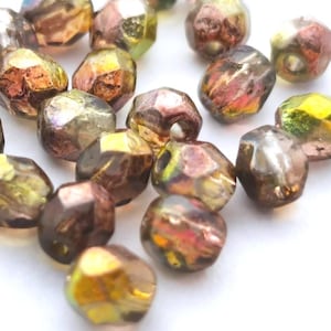 Op de afbeelding: Een verzameling kleine, gefacetteerde glaskralen met een iriserende afwerking in wisselende tinten van metallic brons, olijfgroen en goud. Elke kraal heeft een veelzijdig oppervlak dat licht reflecteert en een glinsterend effect creëert.