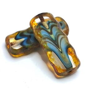 Könnte beinhalten: Zwei rechteckige Glasperlen mit einem wellenförmigen Muster in Blau-, Braun- und Goldtönen. Die Perlen haben ein Loch zum Auffädeln und sind vor einem weißen Hintergrund platziert. Diese Perlen eignen sich für die Schmuckherstellung.