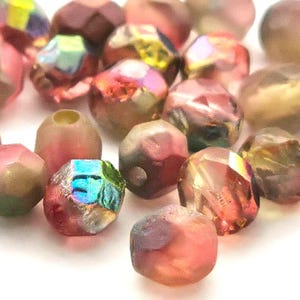 May include: A collection of small, faceted glass beads featuring a mix of translucent pink, muted green, and metallic gold tones. Each bead has a multi-faceted surface with some showing an iridescent, rainbow-like sheen that reflects light.