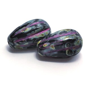 Puede incluir: Dos cuentas en forma de lágrima con un color base oscuro, acentuadas por vetas moradas y tonos iridiscentes. Las cuentas tienen un acabado brillante y probablemente se utilizan para la fabricación de joyas.