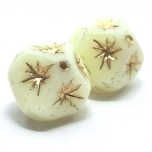 Puede incluir: Dos cuentas de color blanquecino, de forma irregular, con diseños dorados de estrellas. Las cuentas tienen un aspecto ligeramente translúcido y están sobre un fondo blanco. Estas cuentas son adecuadas para la fabricación de joyas.