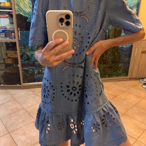 May include: A light blue dress with intricate cut-out floral patterns and short sleeves. The dress features a ruffled hem and a round neckline. The fabric appears to be linen or a similar lightweight material, suitable for warm weather.