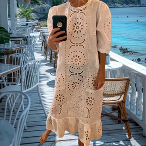 May include: A white, knee-length dress with intricate floral cut-out patterns. The dress features short sleeves and a ruffled hem. The person is holding a black smartphone. The background shows a seaside cafe with white tables and chairs.