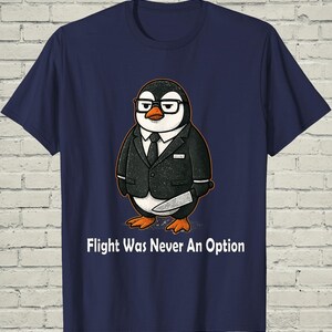 May include: A navy blue short-sleeve t-shirt featuring a graphic of a penguin wearing a black suit, a white shirt, a black tie, and black-rimmed glasses. The penguin holds a knife in one flipper. The text reads: Flight Was Never An Option.