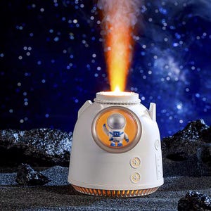 Puede incluir: Humidificador blanco y naranja con temática de astronauta con una pequeña figura de astronauta dentro de una ventana. El humidificador emite una luz naranja brillante desde la parte superior, que se asemeja a un lanzamiento de cohete, y se encuentra sobre una superficie oscura y texturizada.