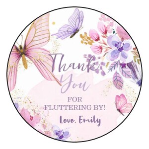 May include: A circular sticker featuring a watercolor design with pink and purple butterflies and floral accents. The text reads Thank You FOR FLUTTERING BY! Love, Emily in a purple script and sans-serif font against a white background.