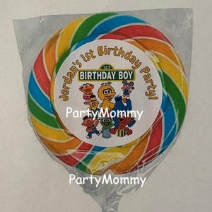 May include: A round, multi-colored swirl lollipop wrapped in clear plastic. A circular sticker on the front features Sesame Street characters and the text: Jordan's 1st Birthday Party! BIRTHDAY BOY. The candy has red, yellow, green, and blue stripes.