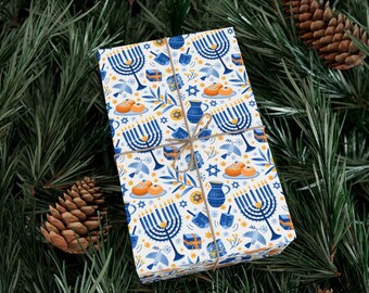 Puede incluir: Un regalo envuelto con un patrón temático de Janucá. El papel de regalo presenta menorás azules, dreidels, Estrellas de David y otros símbolos festivos sobre un fondo blanco. El regalo está atado con hilo y colocado sobre un fondo de agujas de pino y piñas.
