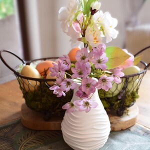 Könnte beinhalten: Eine weiße Vase mit wellenförmigem Design hält rosa und weiße Blüten. Ein Drahtkorb mit Eiern und Moos steht hinter der Vase und bildet einen frühlingshaften Mittelpunkt. Der Korb steht auf einem Holzsockel.