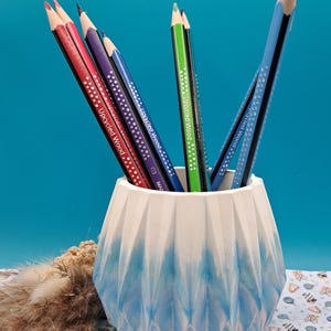 Könnte beinhalten: Eine Sammlung von Farbstiften in einem weiß-blauen Halter mit geometrischem Muster. Die Stifte sind in verschiedenen Farben, darunter Rot, Lila, Grün und Blau, und tragen die Aufschrift "Upcycled Wood". Der Halter hat einen blauen Ombre-Effekt.